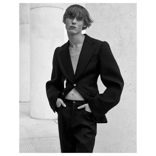 A stylish adult male model in a black suit with an open jacket poses against a neutral wall with architectural columns in a monochrome portrait.