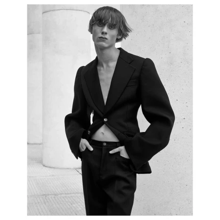 A stylish adult male model in a black suit with an open jacket poses against a neutral wall with architectural columns in a monochrome portrait.