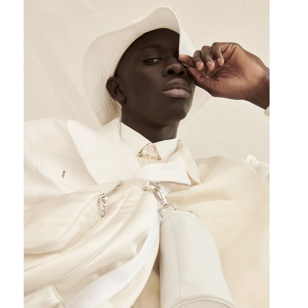 Close-up studio shot of a black man in a white jacket and hat, touching his face, with soft, diffused lighting and a neutral backdrop.