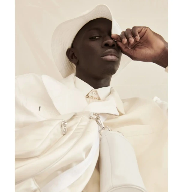 Close-up studio shot of a black man in a white jacket and hat, touching his face, with soft, diffused lighting and a neutral backdrop.