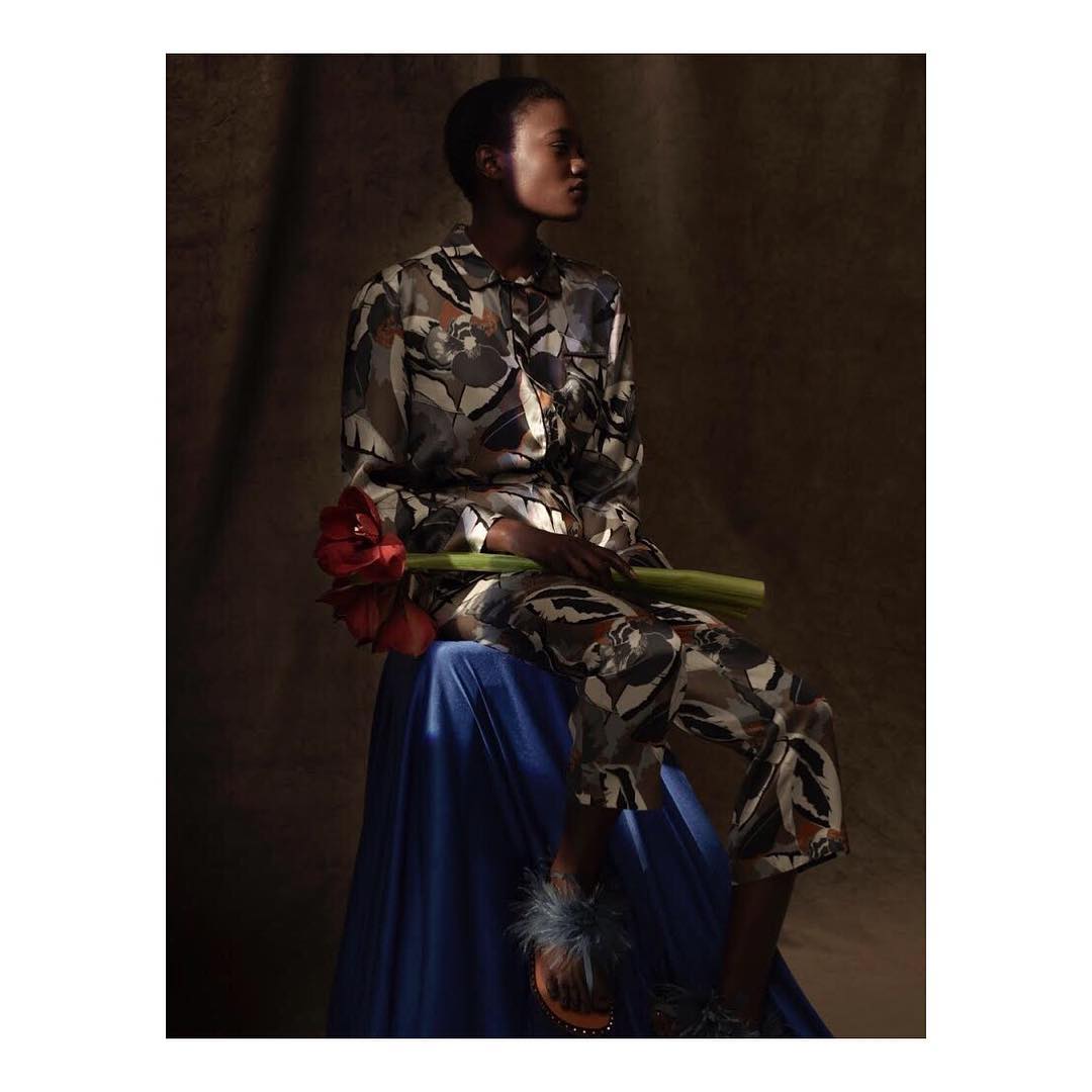 An adult Black woman in patterned matching set sitting with red amaryllis flowers on a blue draped surface in a dark studio.