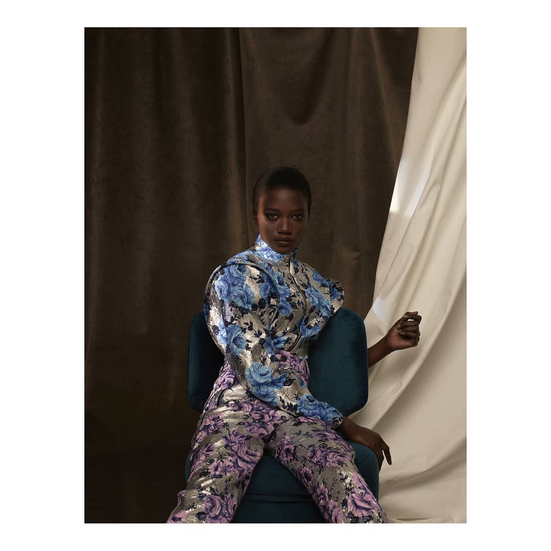 Fashionable woman in floral brocade top and pants seated in teal chair against a draped studio backdrop, eyes forward, elegant pose.