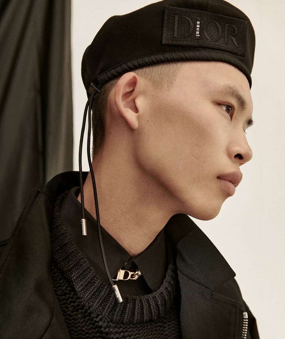 Asian man in a black beret and matching knit sweater and jacket gazes to the right against an off-white backdrop with a dark curtain.