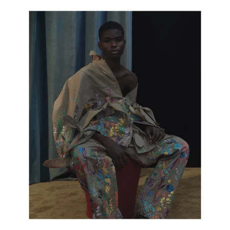 Close-up of a young Black male model in neutral-toned, splattered jacket and pants, seated on a red stool against a black and teal backdrop.