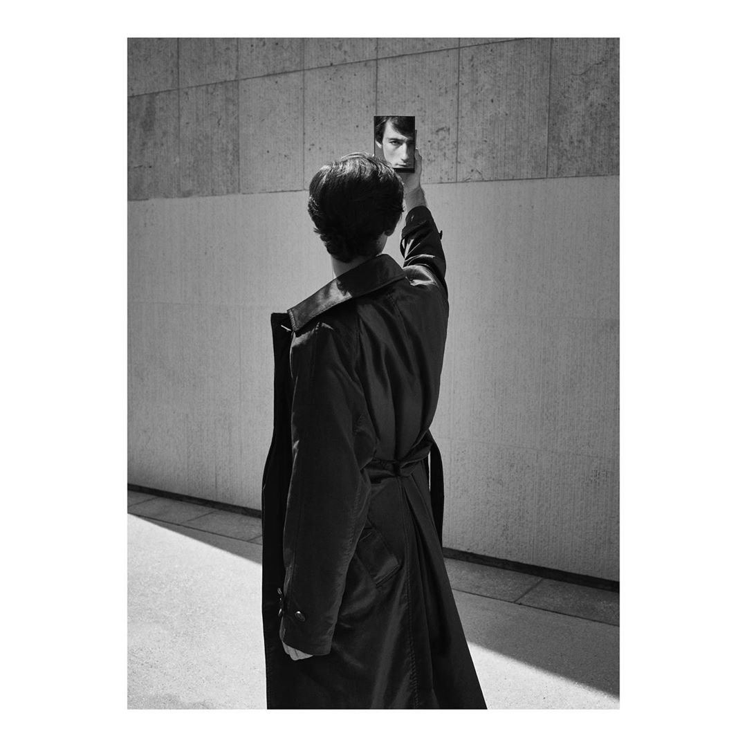 Monochrome portrait of a woman in trench coat holding mirror reflecting a man's face against a concrete wall background.