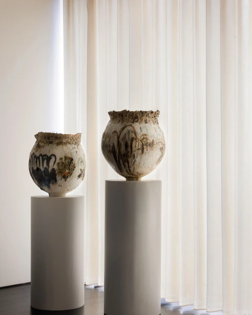 Two decorative vases with textured surfaces sit atop minimalist cylindrical pedestals, set against a backdrop of neutral-toned curtains.