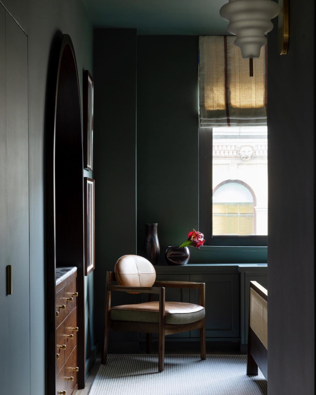 Indoor view of an armchair with flowers, vase, and window. Elegant interior design with natural light and dark green walls.