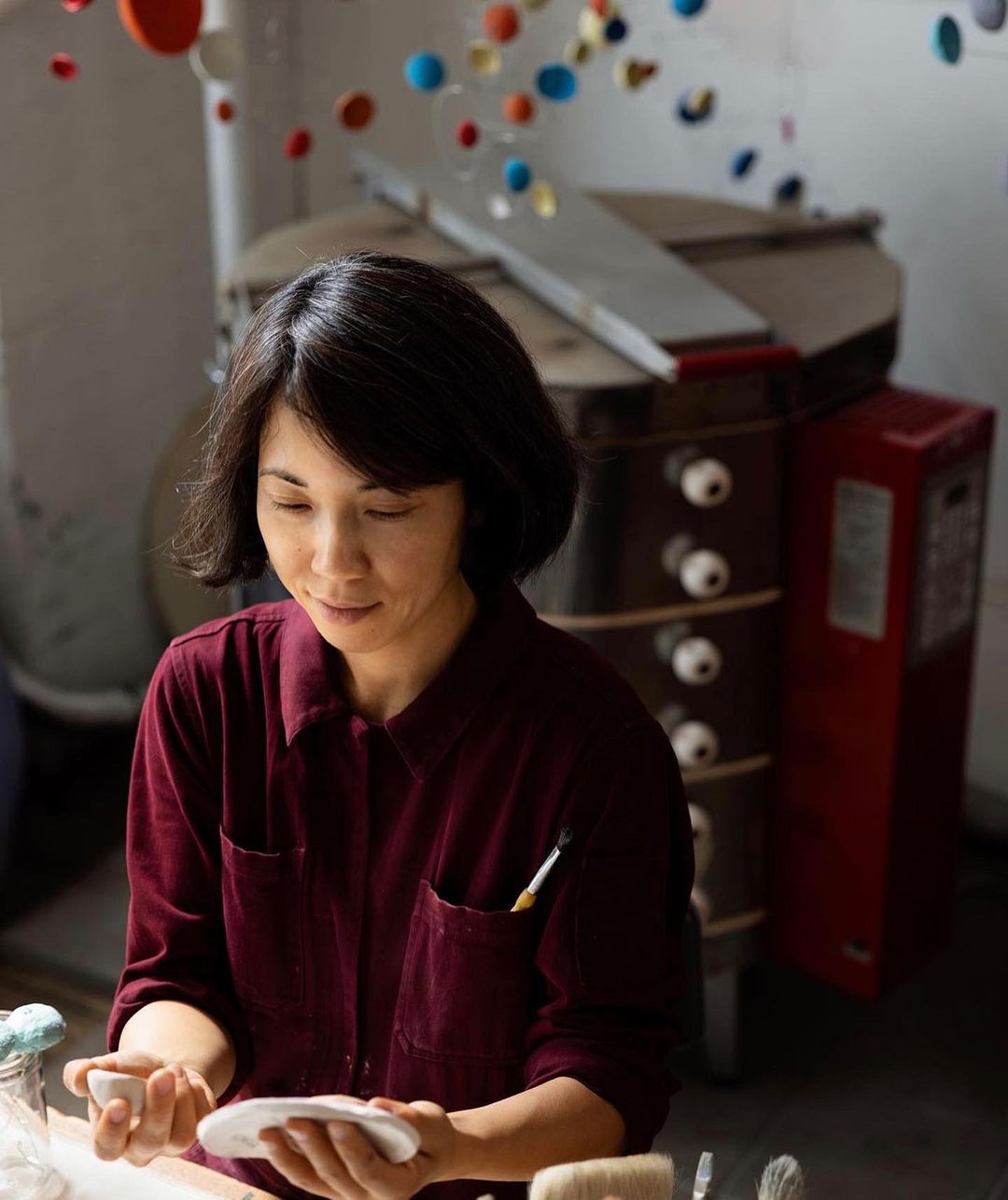 Asian woman crafting a white ceramic piece in a pottery studio with natural light and a kiln in the background, wearing a maroon shirt.