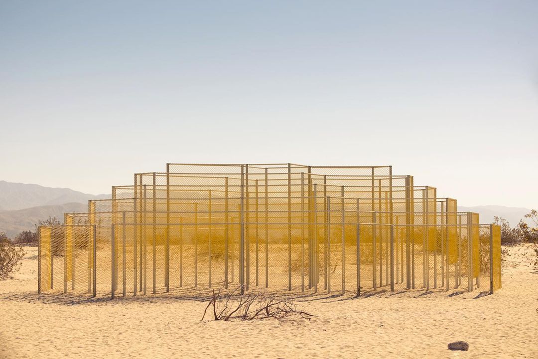 Abstract geometric landscape art installation in the desert with fence-like metal frames, minimalist, set against a clear sky and distant mountains.