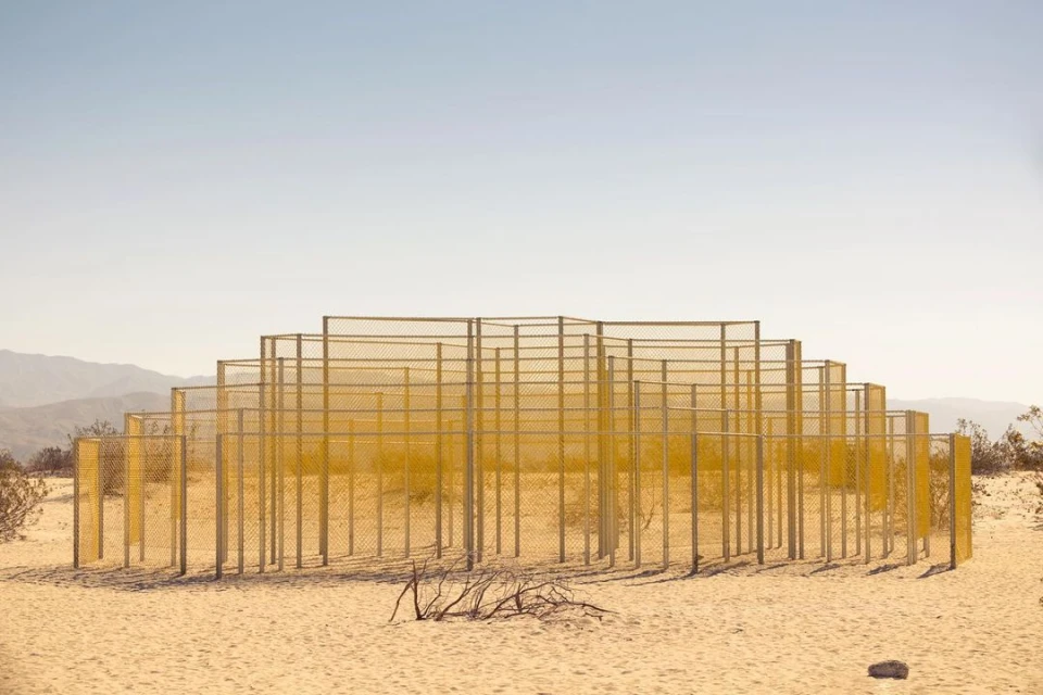 Abstract geometric landscape art installation in the desert with fence-like metal frames, minimalist, set against a clear sky and distant mountains.