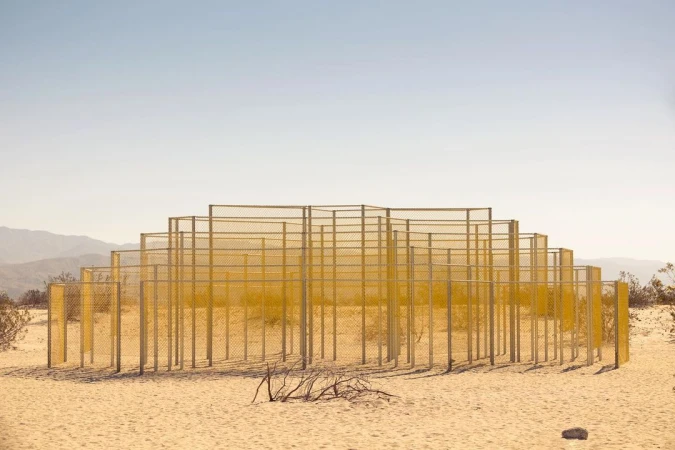 Abstract geometric landscape art installation in the desert with fence-like metal frames, minimalist, set against a clear sky and distant mountains.