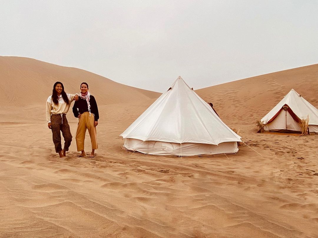 Two women pose in a desert landscape near white canvas tents, under a cloudy sky, with a sand dune backdrop, showcasing a desert camp.