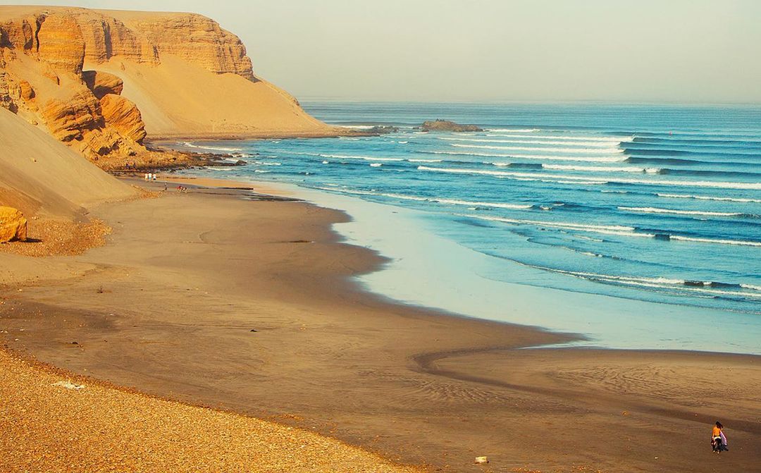 Coastal landscape featuring ocean waves, sandy beach, and a tall, sandy cliff on a sunny day, capturing natural beauty and tranquility.