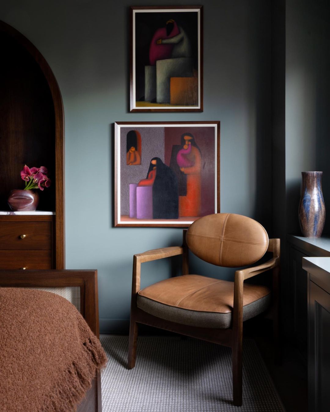 Interior design photograph: A modern armchair with a light-brown leather seat and light gray rug are below framed art on a dark blue wall.