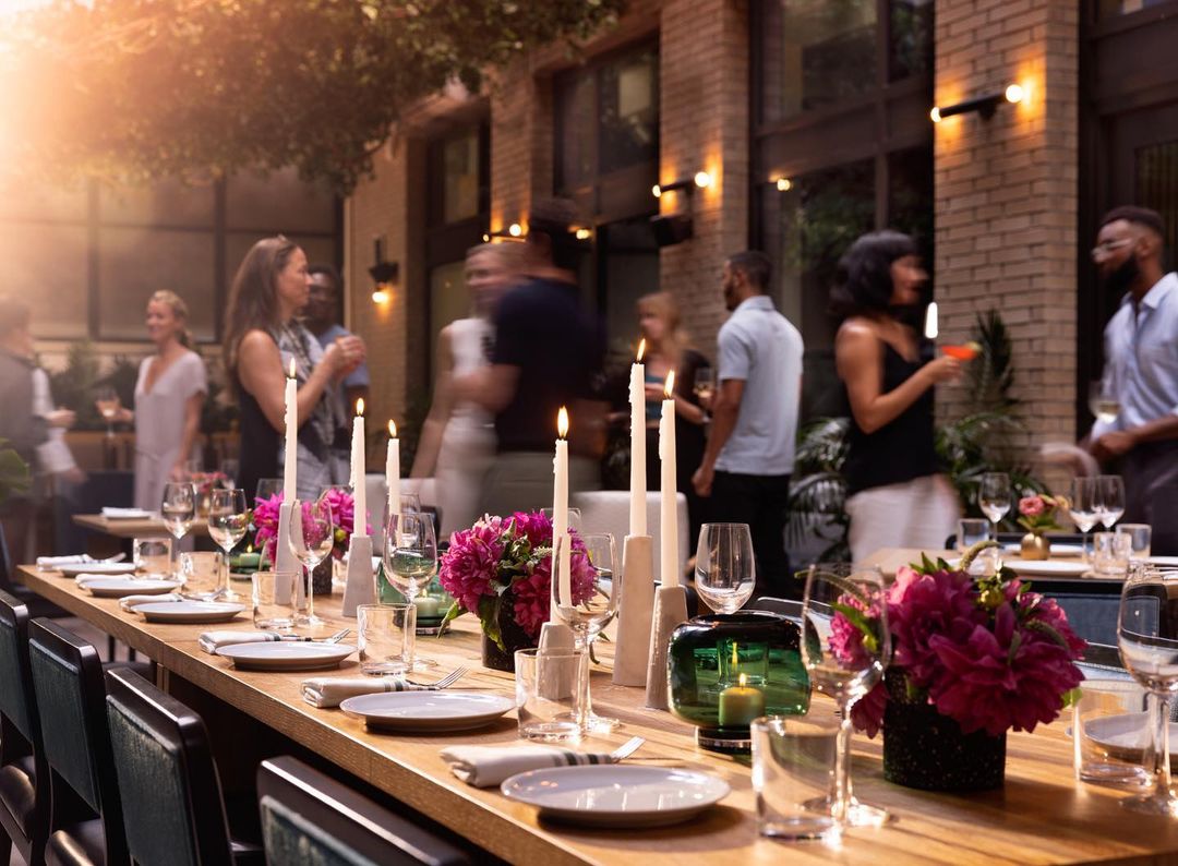 Outdoor dining event with long table, candles, floral arrangements, set plates and glasses, and a group of people mingling around.