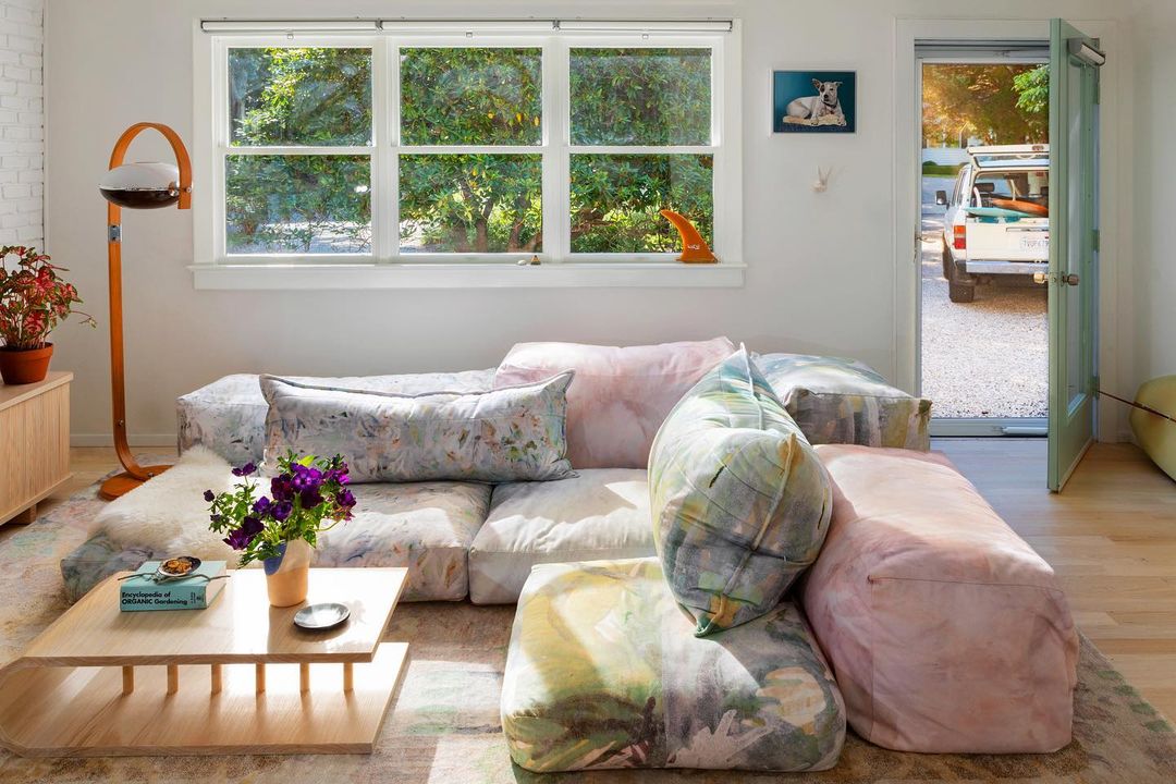 Living room with a light wood coffee table, a modular sofa with pastel cushions, and a window overlooking lush green foliage.