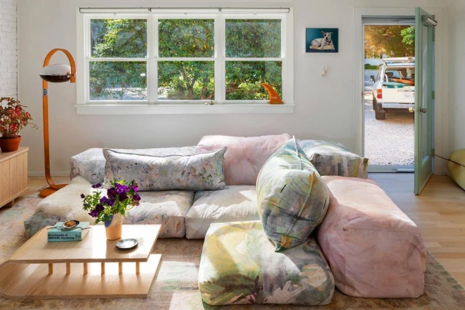 Living room with a light wood coffee table, a modular sofa with pastel cushions, and a window overlooking lush green foliage.
