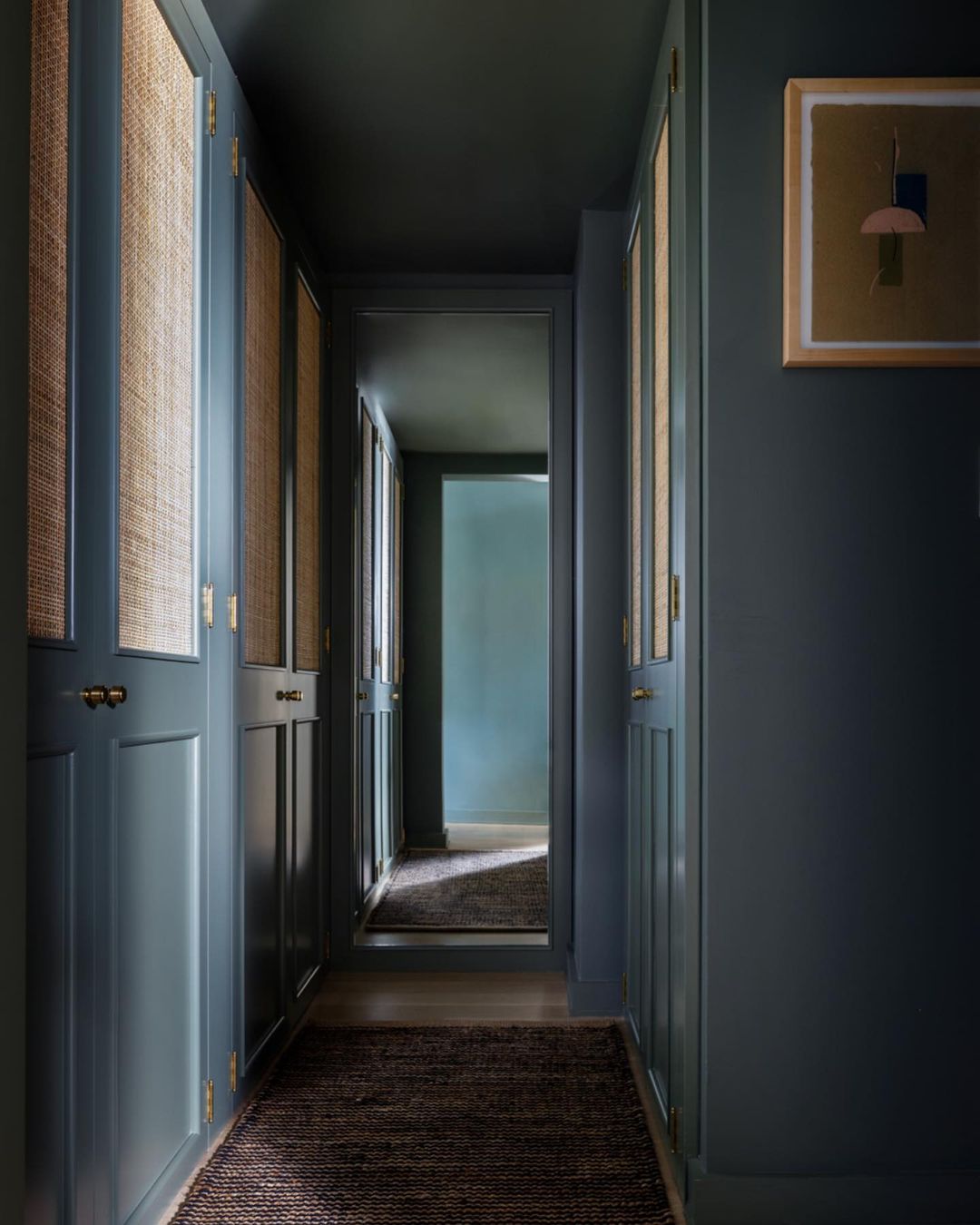 Interior view of a hallway with dark blue walls, wood-paneled doors, wicker screens, an abstract painting, and a jute rug.