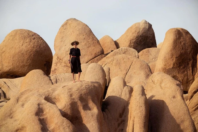 Woman in a black dress and hat standing atop large rounded desert rock formations under an open sky