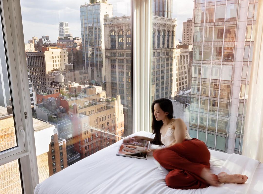 Asian woman in a white top and red pants sitting on a bed reading a magazine with a skyscraper-filled cityscape background.