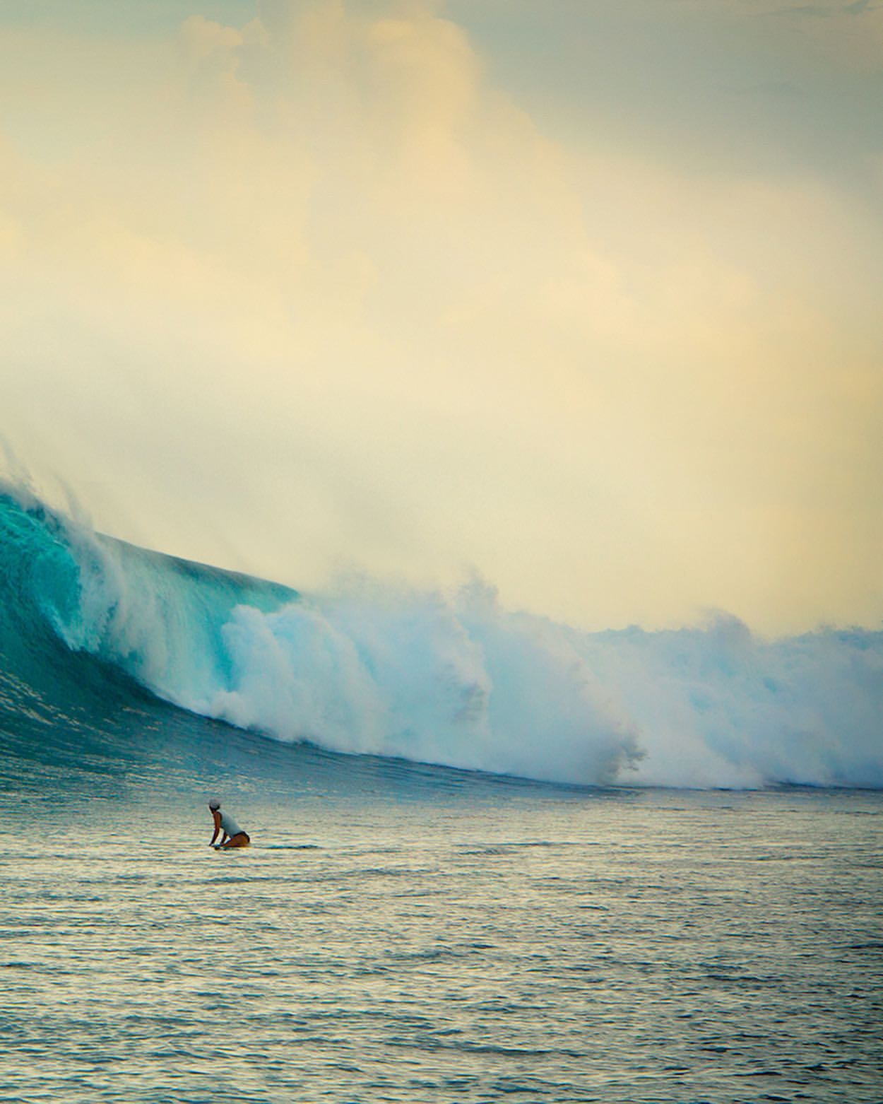 Surfer paddles on board towards massive wave with turquoise water cresting under a pale cloudy sky, creating a dramatic ocean scene.