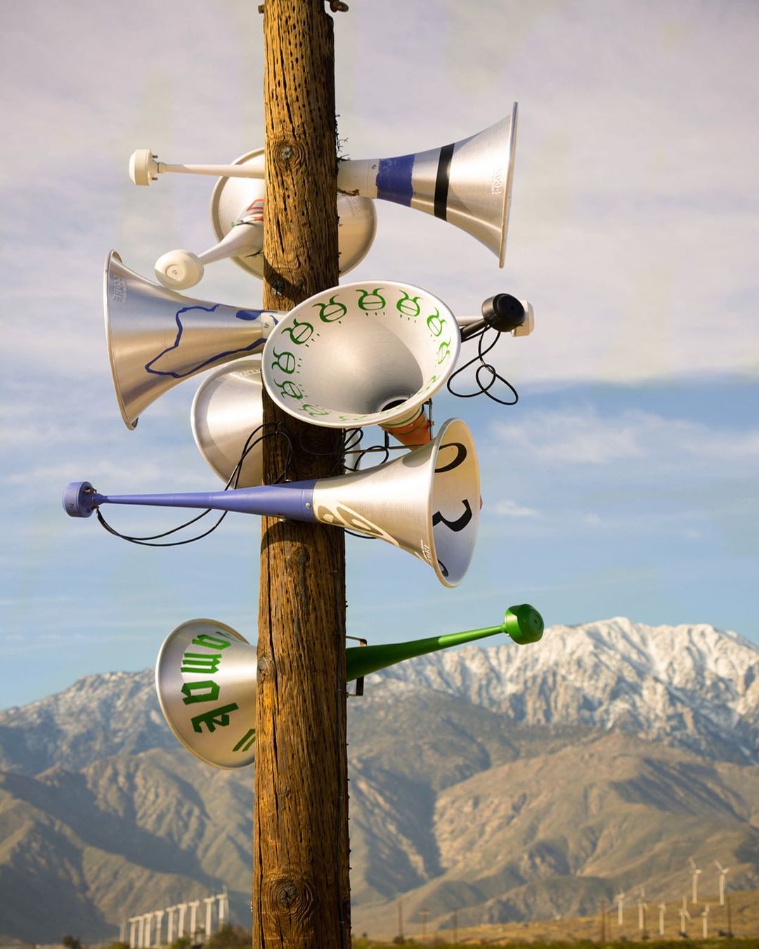 Multiple silver megaphone-shaped loudspeakers attached to a wooden pole in a desert landscape with blue sky and mountains.