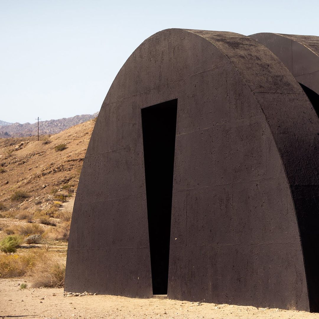 Modern, dark architectural structure in desert landscape featuring repeating archways creating a tunnel or passageway in the daytime.