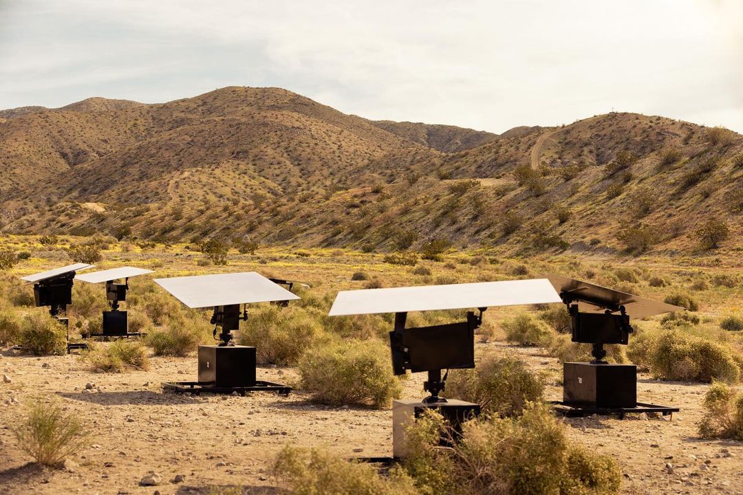 Several solar panels stand in a desert environment with mountains in the background. They are positioned among desert scrub.