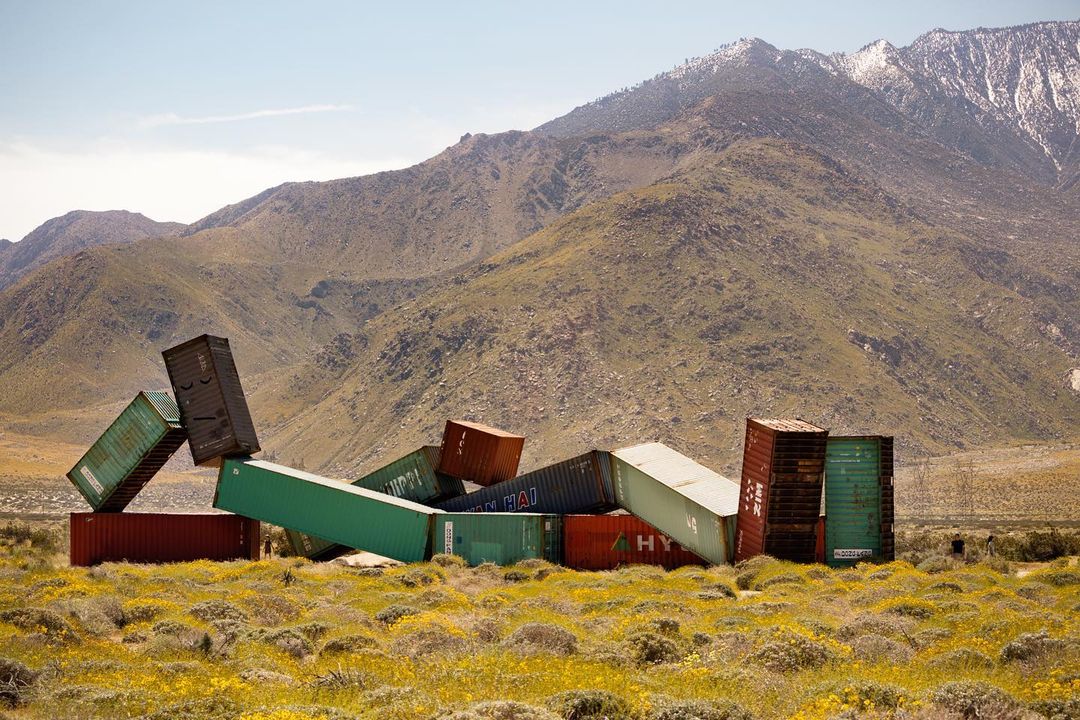 A surreal desert landscape features a colorful pile of shipping containers against a mountain backdrop, creating a unique visual contrast.