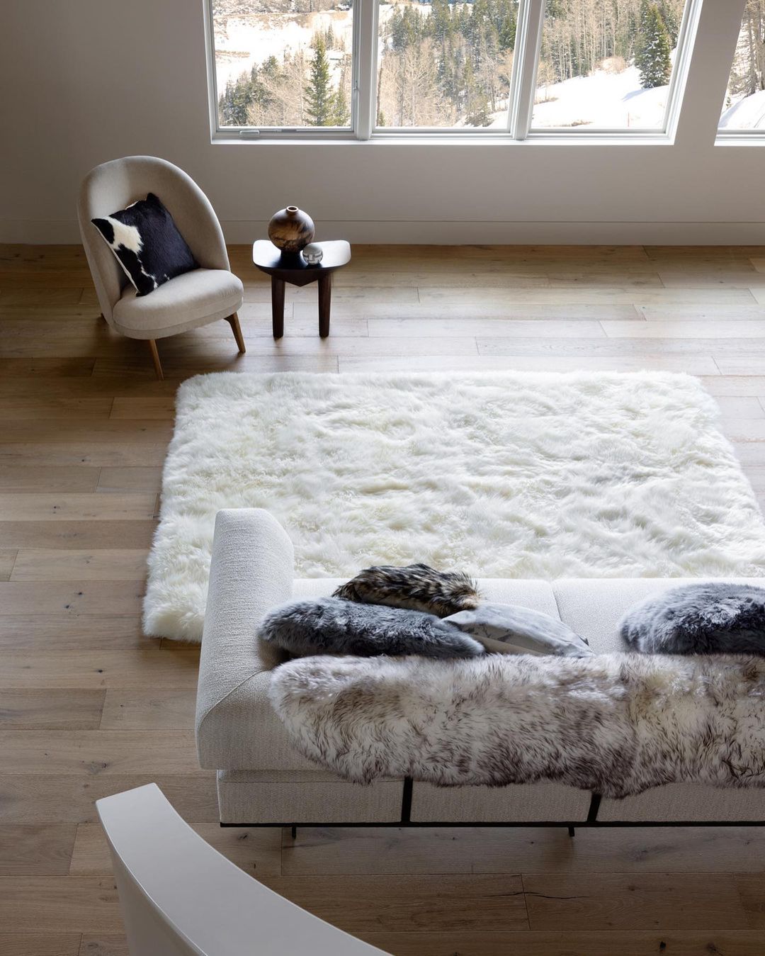 Living room interior featuring a white sofa with fur throws, a shag rug, a modern chair, and a window view of a snow-covered mountain.