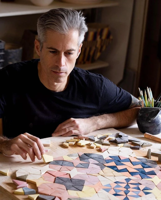 Adult man creates mosaic artwork with colored tiles on a wooden table inside a bright, natural light-filled studio. Close-up shot.