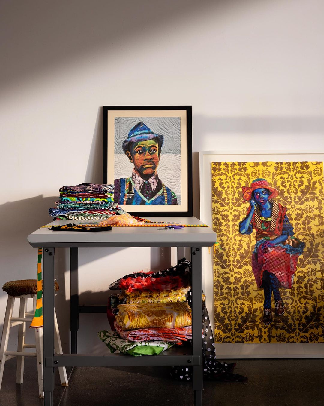 Interior shot features colorful artwork, stacks of patterned fabric, and a gray worktable against a white wall, complemented by a stool.