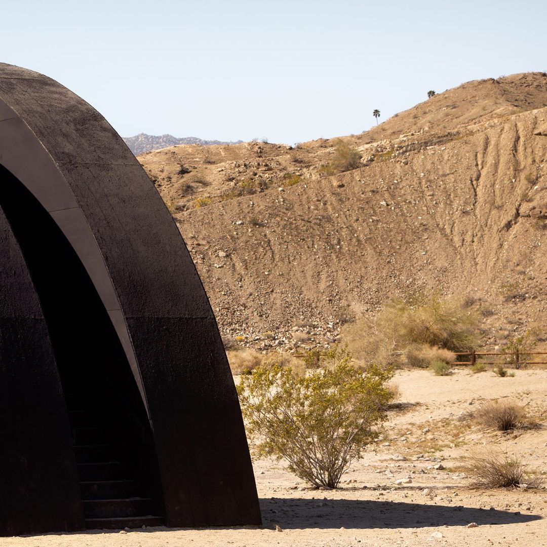 Dark arched structure with steps leading down in a desert landscape with arid hills under a clear sky.