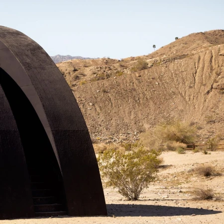 Dark arched structure with steps leading down in a desert landscape with arid hills under a clear sky.