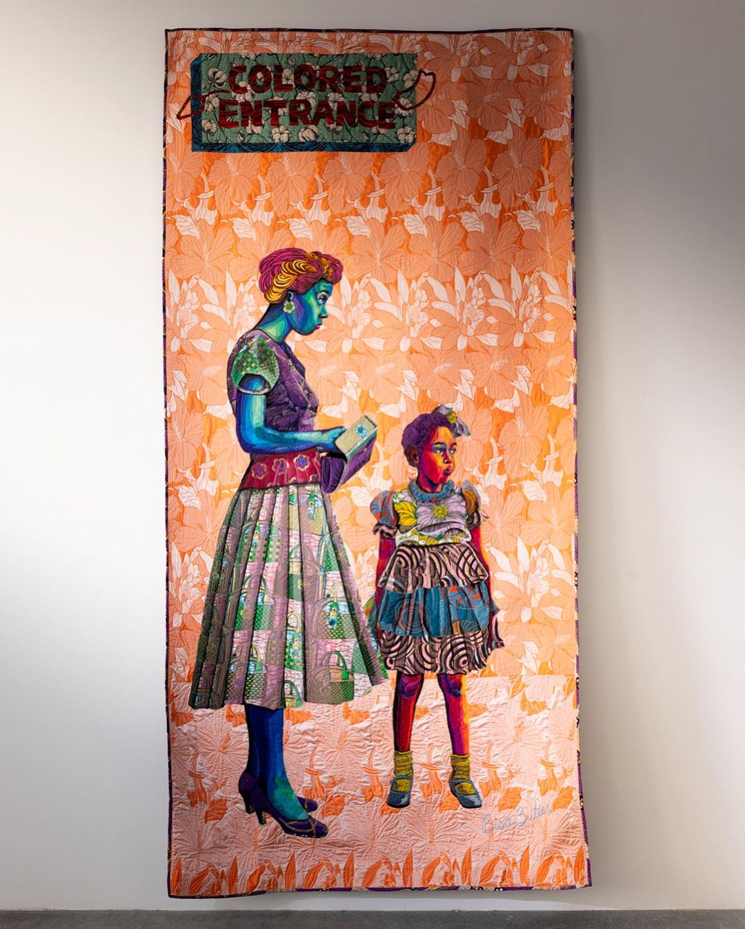 A mixed-media artwork on fabric features an African-American woman and child in a vintage style standing under a "Colored Entrance" sign.