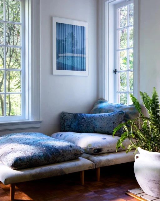 Reading nook with patterned cushions by the window, indoor plants and art piece hanging on the wall in a light-filled room.