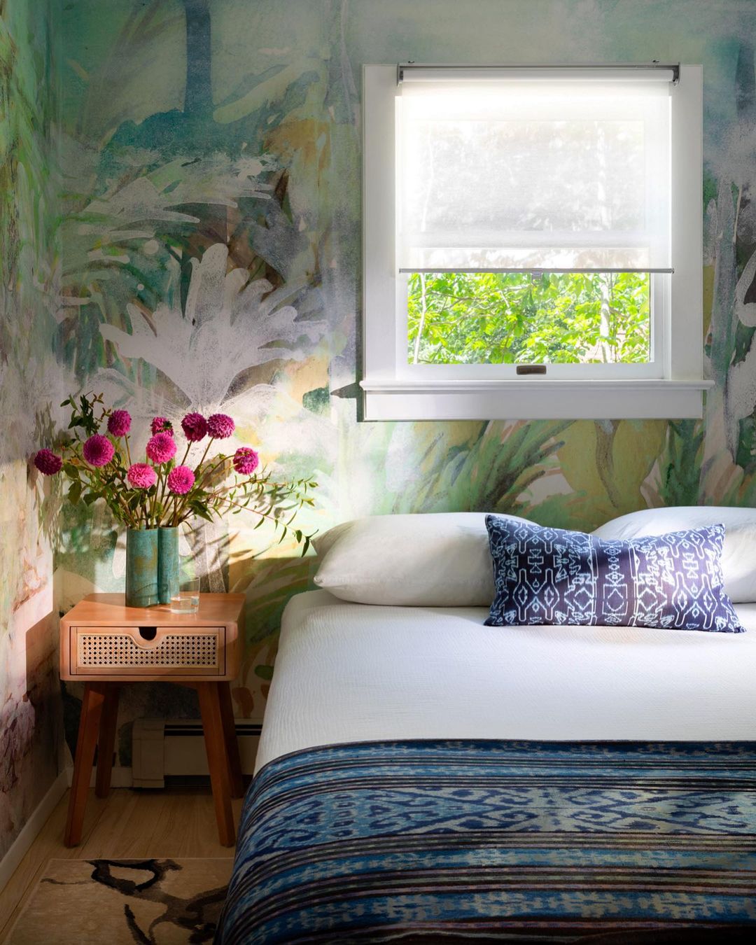 Bedroom interior featuring a bed with patterned blue blanket and pillow, a wooden nightstand with flowers, and botanical wallpaper.