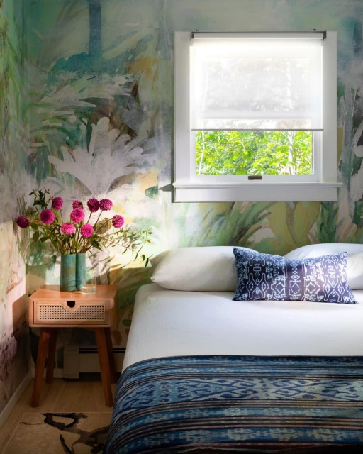Bedroom interior featuring a bed with patterned blue blanket and pillow, a wooden nightstand with flowers, and botanical wallpaper.