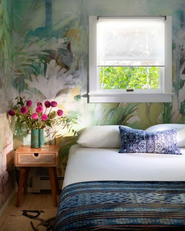 Bedroom interior featuring a bed with patterned blue blanket and pillow, a wooden nightstand with flowers, and botanical wallpaper.