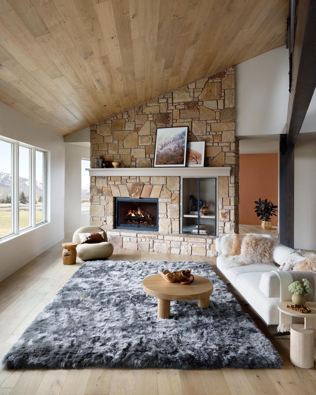 Living room interior with a stone fireplace, light wood flooring, a gray shag rug, and a white sofa under a vaulted wood ceiling.