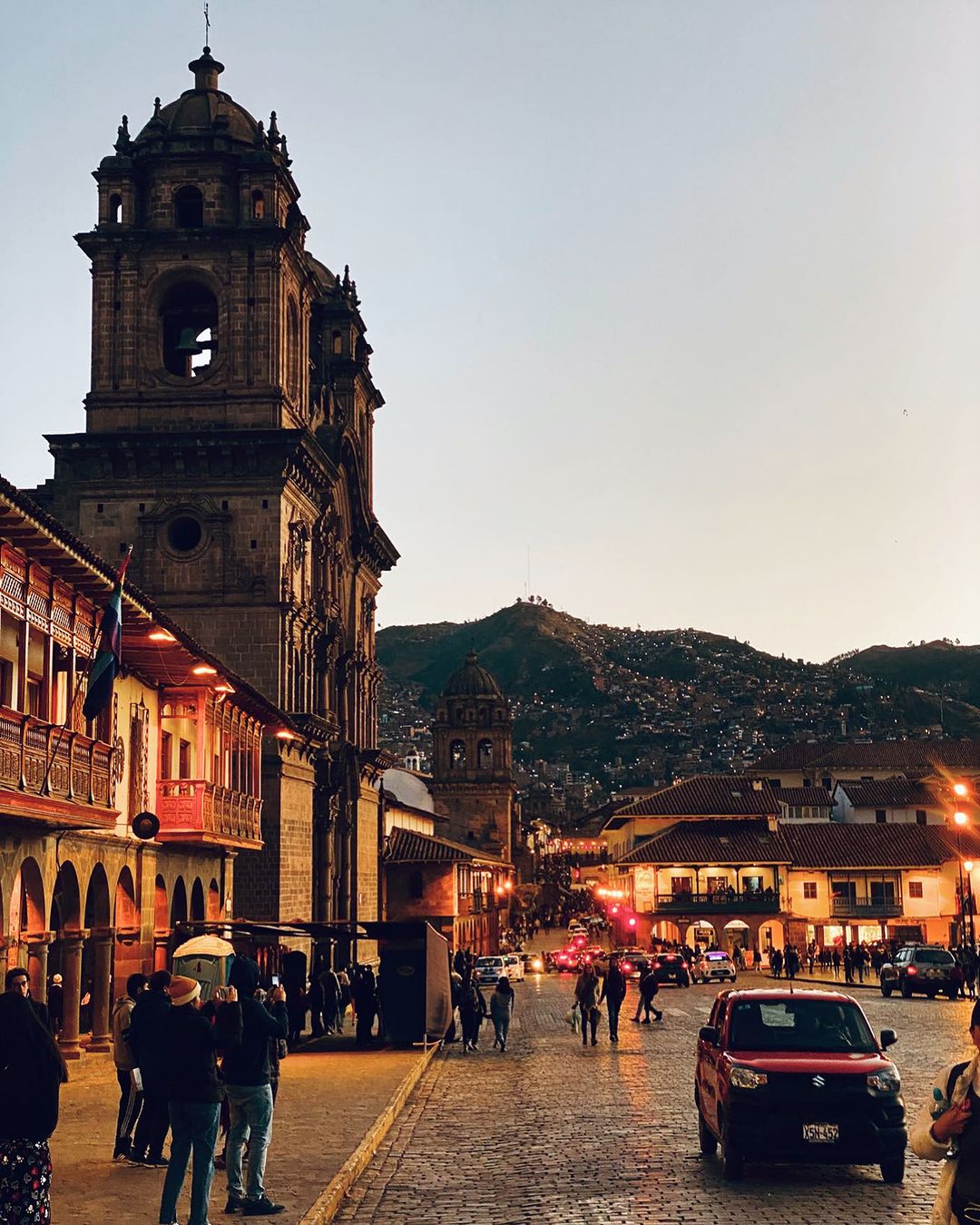 Scenic view of Cusco, Peru featuring an ornate cathedral and surrounding buildings under a twilight sky with pedestrian and vehicle traffic.
