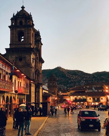 Scenic view of Cusco, Peru featuring an ornate cathedral and surrounding buildings under a twilight sky with pedestrian and vehicle traffic.