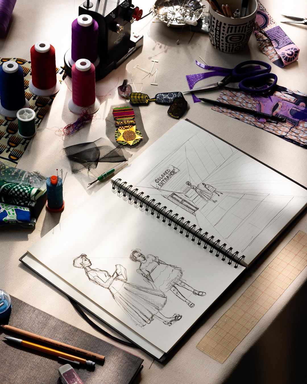 Open sketchbook on a craft table showing fashion design sketches, surrounded by threads, tools, and fabric scraps in a studio setting.