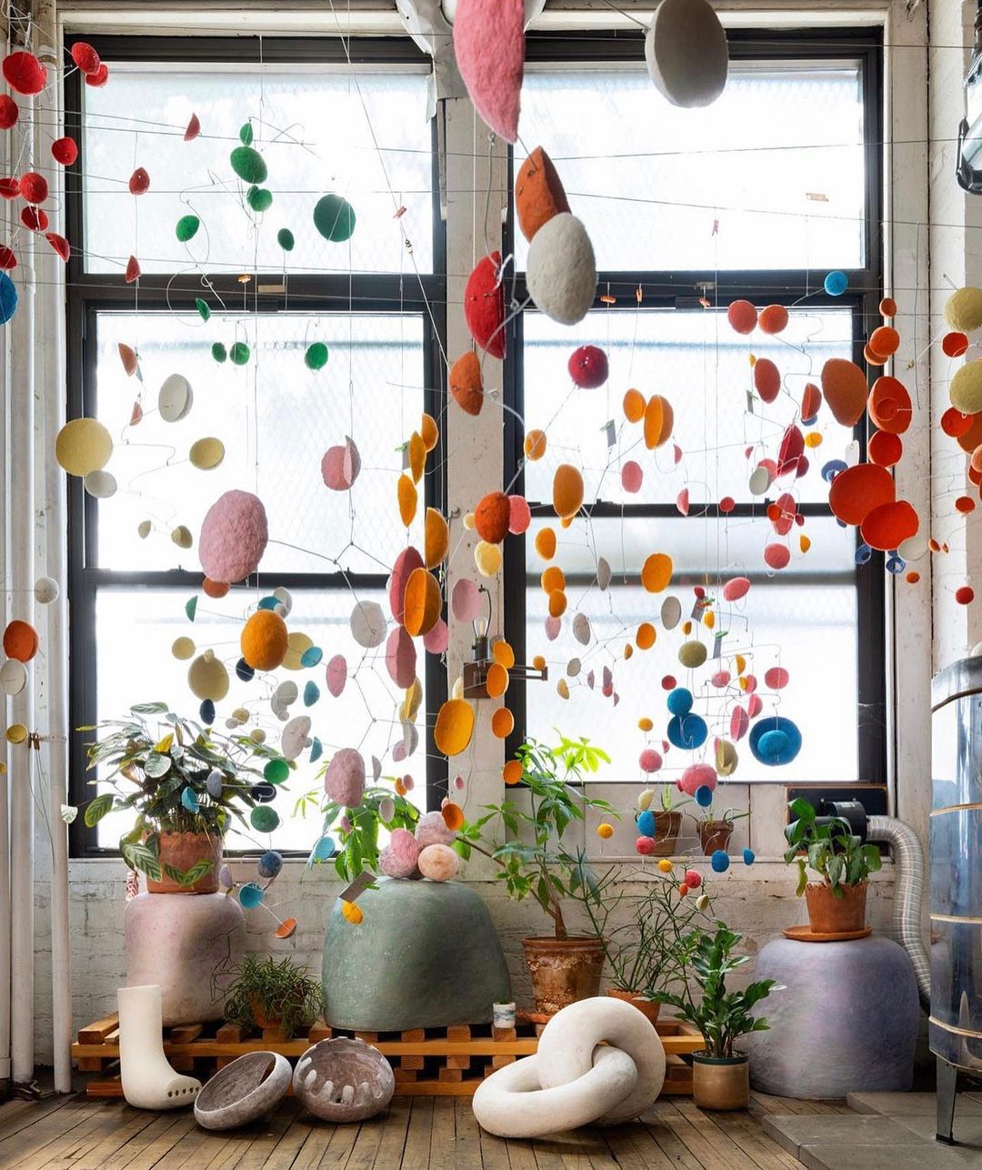 Indoor shot of colorful hanging mobile art installation near a window, with several potted plants, white decorative objects, and wood pallet.