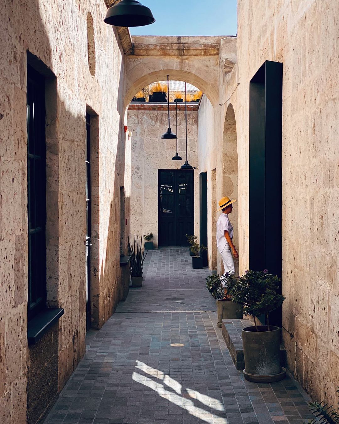 Woman in white suit and straw hat stands in stone archway of a colonial-style building, plants and shadows, travel photography.