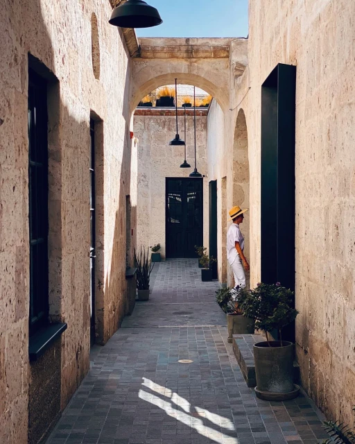 Woman in white suit and straw hat stands in stone archway of a colonial-style building, plants and shadows, travel photography.