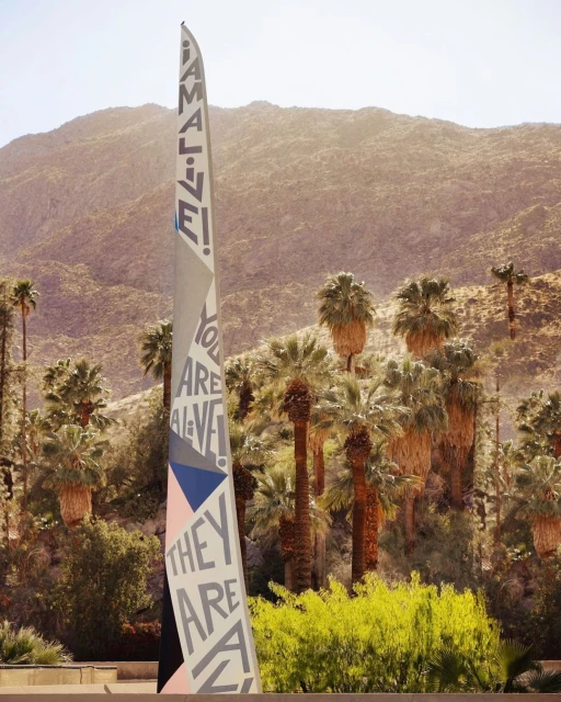 Art installation with 'I AM ALIVE!' message, set against palm trees and mountains in a sunlit, arid landscape, creating a vibrant contrast.
