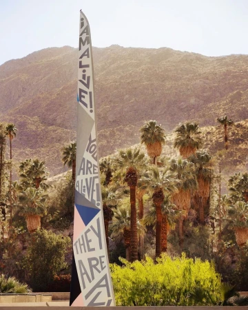 Art installation with 'I AM ALIVE!' message, set against palm trees and mountains in a sunlit, arid landscape, creating a vibrant contrast.