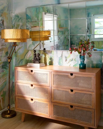 Interior view of a light-colored wooden dresser with cane drawers, decorated with a lamp, vase, and sculpture against a patterned wallpaper.