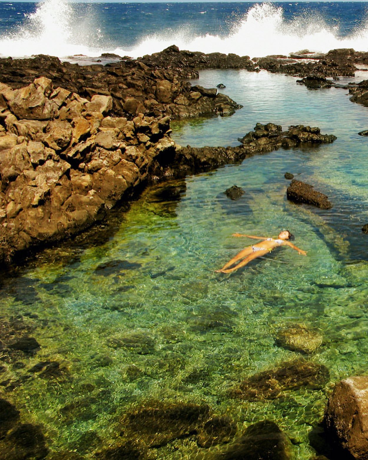 Woman floating in clear turquoise ocean tide pool surrounded by black volcanic rock formations and ocean waves crashing in the background.